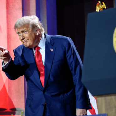 Donald Trump, vestindo terno azul e gravata vermelha, aponta com o dedo indicador durante um discurso. Ao fundo, colunas iluminadas em vermelho e azul e o selo presidencial dos Estados Unidos estão visíveis, reforçando o cenário oficial.