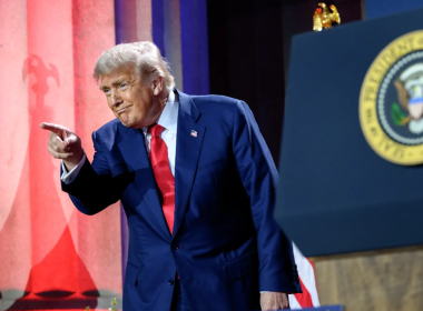 Donald Trump, vestindo terno azul e gravata vermelha, aponta com o dedo indicador durante um discurso. Ao fundo, colunas iluminadas em vermelho e azul e o selo presidencial dos Estados Unidos estão visíveis, reforçando o cenário oficial.