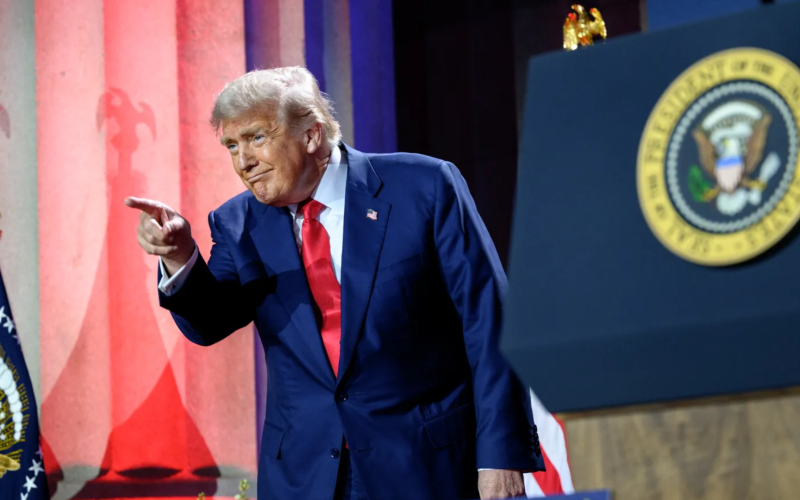 Donald Trump, vestindo terno azul e gravata vermelha, aponta com o dedo indicador durante um discurso. Ao fundo, colunas iluminadas em vermelho e azul e o selo presidencial dos Estados Unidos estão visíveis, reforçando o cenário oficial.