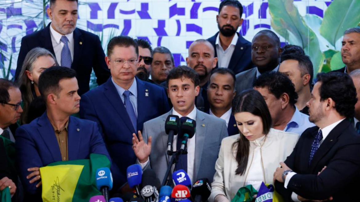 Deputado Nikolas Ferreira discursa em coletiva de imprensa cercado por aliados e apoiadores, diante de microfones de diversas emissoras, em evento político com bandeiras do Brasil.