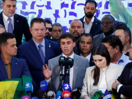 Deputado Nikolas Ferreira discursa em coletiva de imprensa cercado por aliados e apoiadores, diante de microfones de diversas emissoras, em evento político com bandeiras do Brasil.