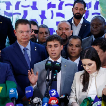 Deputado Nikolas Ferreira discursa em coletiva de imprensa cercado por aliados e apoiadores, diante de microfones de diversas emissoras, em evento político com bandeiras do Brasil.