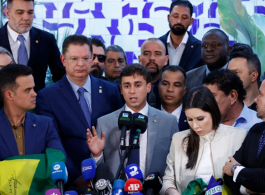 Deputado Nikolas Ferreira discursa em coletiva de imprensa cercado por aliados e apoiadores, diante de microfones de diversas emissoras, em evento político com bandeiras do Brasil.