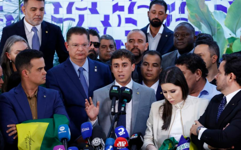 Deputado Nikolas Ferreira discursa em coletiva de imprensa cercado por aliados e apoiadores, diante de microfones de diversas emissoras, em evento político com bandeiras do Brasil.