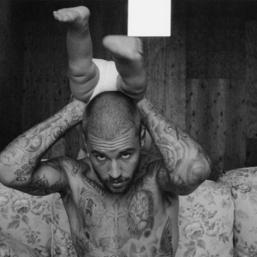 Justin Bieber aparece tatuado, sem camisa, sentado em um sofá floral, segura um bebê de ponta-cabeça apoiado sobre sua cabeça, com as pernas do bebê direcionadas para cima. A imagem está em preto e branco e tem um estilo artístico e intimista.