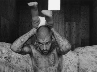 Justin Bieber aparece tatuado, sem camisa, sentado em um sofá floral, segura um bebê de ponta-cabeça apoiado sobre sua cabeça, com as pernas do bebê direcionadas para cima. A imagem está em preto e branco e tem um estilo artístico e intimista.