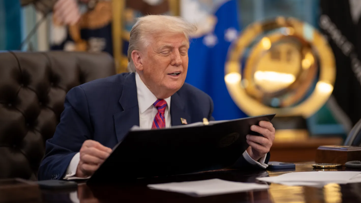Donald Trump, sentado em uma mesa no Salão Oval da Casa Branca, veste terno azul e gravata vermelha enquanto segura uma pasta preta. Ele está com os olhos semicerrados, aparentando falar ou reagir a algo. Ao fundo, há bandeiras oficiais e um grande emblema dourado da Força Espacial dos EUA. Sobre a mesa, há papéis, um telefone e um porta-canetas.