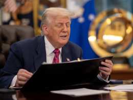 Donald Trump, sentado em uma mesa no Salão Oval da Casa Branca, veste terno azul e gravata vermelha enquanto segura uma pasta preta. Ele está com os olhos semicerrados, aparentando falar ou reagir a algo. Ao fundo, há bandeiras oficiais e um grande emblema dourado da Força Espacial dos EUA. Sobre a mesa, há papéis, um telefone e um porta-canetas.