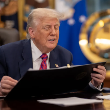Donald Trump, sentado em uma mesa no Salão Oval da Casa Branca, veste terno azul e gravata vermelha enquanto segura uma pasta preta. Ele está com os olhos semicerrados, aparentando falar ou reagir a algo. Ao fundo, há bandeiras oficiais e um grande emblema dourado da Força Espacial dos EUA. Sobre a mesa, há papéis, um telefone e um porta-canetas.