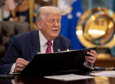 Donald Trump, sentado em uma mesa no Salão Oval da Casa Branca, veste terno azul e gravata vermelha enquanto segura uma pasta preta. Ele está com os olhos semicerrados, aparentando falar ou reagir a algo. Ao fundo, há bandeiras oficiais e um grande emblema dourado da Força Espacial dos EUA. Sobre a mesa, há papéis, um telefone e um porta-canetas.
