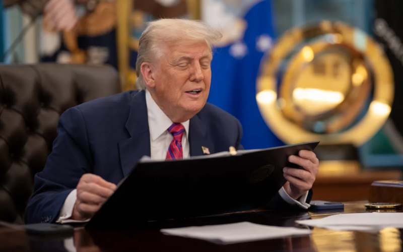Donald Trump, sentado em uma mesa no Salão Oval da Casa Branca, veste terno azul e gravata vermelha enquanto segura uma pasta preta. Ele está com os olhos semicerrados, aparentando falar ou reagir a algo. Ao fundo, há bandeiras oficiais e um grande emblema dourado da Força Espacial dos EUA. Sobre a mesa, há papéis, um telefone e um porta-canetas.