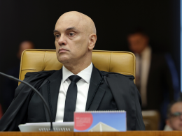 Ministro Alexandre de Moraes sentado na cadeira do plenário do STF, usando toga preta e gravata, com expressão séria durante sessão do Supremo Tribunal Federal. Ao fundo, pessoas desfocadas acompanham a sessão.