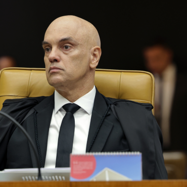 Ministro Alexandre de Moraes sentado na cadeira do plenário do STF, usando toga preta e gravata, com expressão séria durante sessão do Supremo Tribunal Federal. Ao fundo, pessoas desfocadas acompanham a sessão.