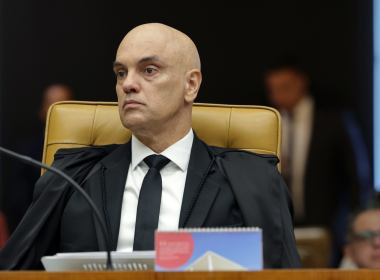 Ministro Alexandre de Moraes sentado na cadeira do plenário do STF, usando toga preta e gravata, com expressão séria durante sessão do Supremo Tribunal Federal. Ao fundo, pessoas desfocadas acompanham a sessão.