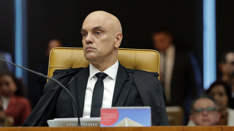 Ministro Alexandre de Moraes sentado na cadeira do plenário do STF, usando toga preta e gravata, com expressão séria durante sessão do Supremo Tribunal Federal. Ao fundo, pessoas desfocadas acompanham a sessão.