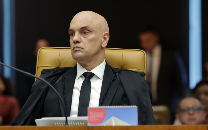 Ministro Alexandre de Moraes sentado na cadeira do plenário do STF, usando toga preta e gravata, com expressão séria durante sessão do Supremo Tribunal Federal. Ao fundo, pessoas desfocadas acompanham a sessão.