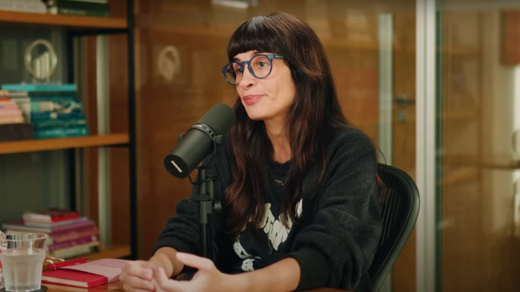 Camila Fremder sentada em estúdio de podcast, usando óculos azuis e moletom preto, falando ao microfone com expressão séria. Ao fundo, estante com livros coloridos e parede de vidro.