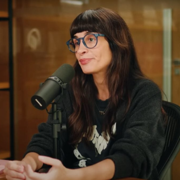 Camila Fremder sentada em estúdio de podcast, usando óculos azuis e moletom preto, falando ao microfone com expressão séria. Ao fundo, estante com livros coloridos e parede de vidro.