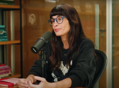 Camila Fremder sentada em estúdio de podcast, usando óculos azuis e moletom preto, falando ao microfone com expressão séria. Ao fundo, estante com livros coloridos e parede de vidro.