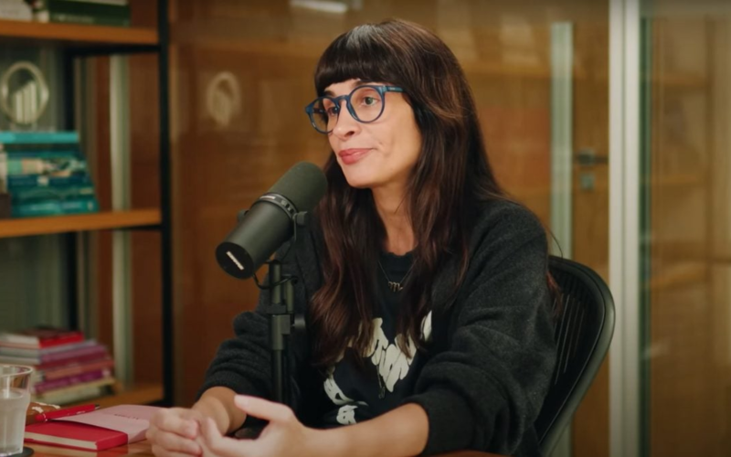 Camila Fremder sentada em estúdio de podcast, usando óculos azuis e moletom preto, falando ao microfone com expressão séria. Ao fundo, estante com livros coloridos e parede de vidro.