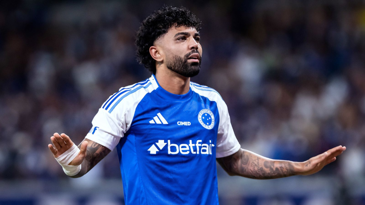 Gabigol com uniforme do Cruzeiro durante partida, com expressão séria e braços estendidos, em estádio com torcida ao fundo.