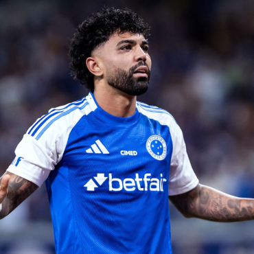 Gabigol com uniforme do Cruzeiro durante partida, com expressão séria e braços estendidos, em estádio com torcida ao fundo.