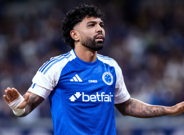 Gabigol com uniforme do Cruzeiro durante partida, com expressão séria e braços estendidos, em estádio com torcida ao fundo.