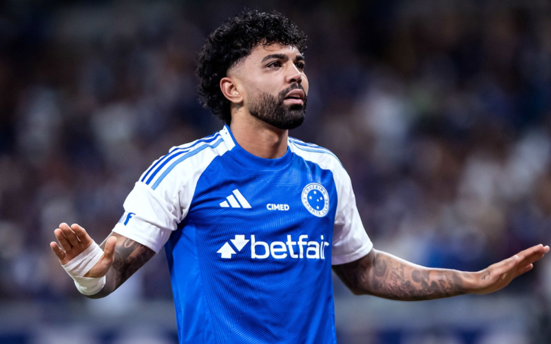 Gabigol com uniforme do Cruzeiro durante partida, com expressão séria e braços estendidos, em estádio com torcida ao fundo.